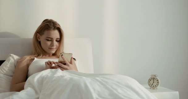 Fiatal csinos szőke lány szép mosollyal boldogan sms-ezik, csevegnek és böngésznek a mobiltelefonon, miközben pihen az ágyban. 4k. — Stock videók