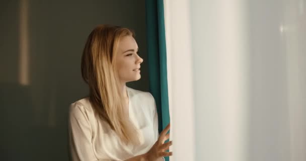 실크 가운을 입은 예쁜 금발의 젊은 여성 이 창문으로 오는 아침입니다. 사이드 초상화. 4k. — 비디오