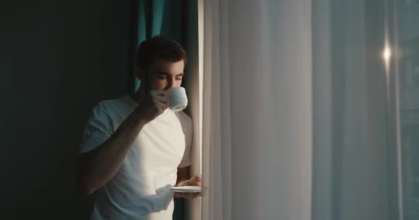 Привлекательный мужчина с темной бородой и волосами наслаждается первой чашкой кофе, глядя на восход солнца через окно. 4k кадров. — стоковое видео