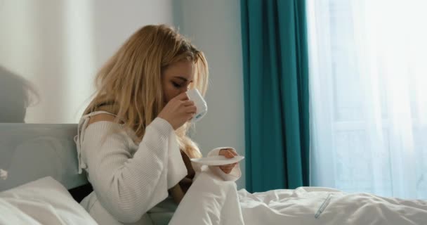 Retrato lateral de la mujer enferma con cabello rubio y cuello envuelto con pañuelo bebiendo taza de té caliente mientras descansa en la cama. Imágenes de 4k. — Vídeo de stock