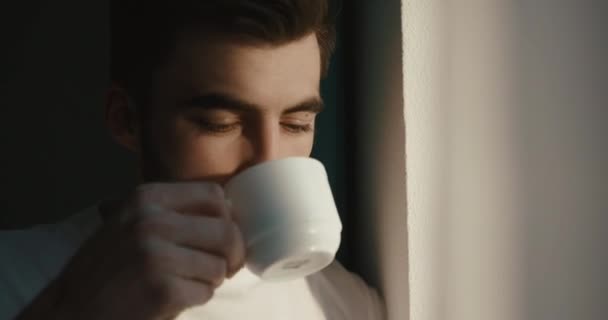 Close-up portret van de vredige man die de eerste kop koffie drinkt en geniet van de zonsopgang door het raam. 4k. — Stockvideo