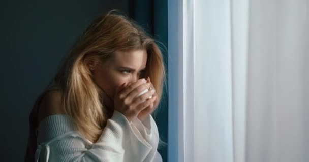 Mulher loira atraente com gripe está bebendo chá para se sentir melhor e olhando pela janela. Imagens de 4K. Retrato lateral. — Vídeo de Stock