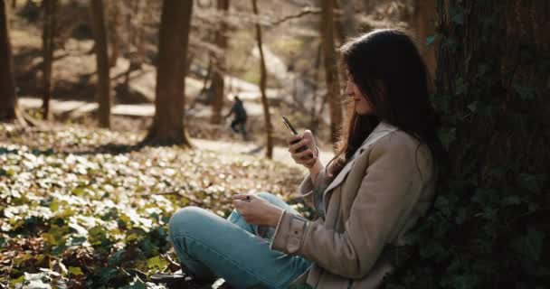 Herfst lente vibes. Charmante jonge vrouw leest iets in haar smartphone zittend in het park — Stockvideo