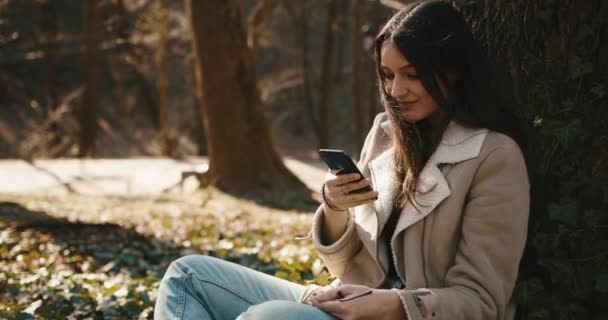 Herfst lente vibes. Charmante jonge vrouw leest iets in haar smartphone zittend in het park — Stockvideo