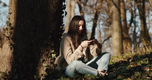 Autumn spring vibes. Angin meniup rambut wanita sementara ia membaca sesuatu di smartphone duduk di taman — Stok Video