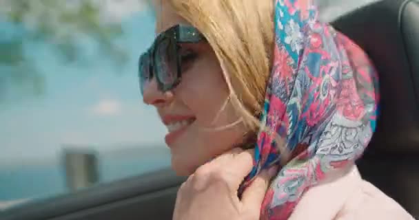 Viaggiare in cabriolet. Donna bionda alla moda sorridente in occhiali da sole e sciarpa sulla testa. Si sta godendo il tempo soleggiato ventoso. 4k filmati. — Video Stock