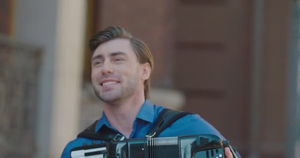 Portrét mladého pohledného muže s hezkým úsměvem hrající na harmoniku na dveřích. Pouliční muzikant. Záběry 4k. — Stock video
