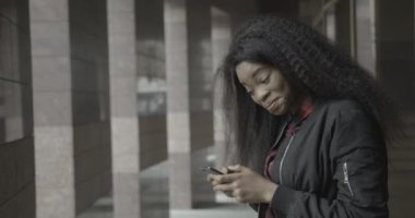 Çekici, mutlu Afrikalı genç kız dışarıda cep telefonuyla mesajlaşırken, sohbet ederken ve dolaşırken gülüyor ve gülümsüyor. 4k görüntü.