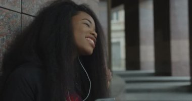 Yan portre. Oldukça duygusal bir Afrikalı kız binaya yaslanırken kulaklıkla müzik dinlemekten zevk alıyor. 4k görüntü.