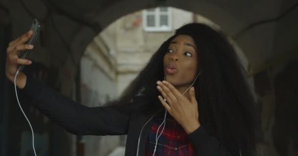 La jeune fille africaine attrayante prend selfies drôles via le téléphone mobile. Elle envoie des baisers d'air, montrant la langue et faisant visage surpris en plein air. — Video