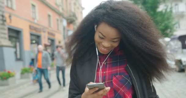 Güzel gülüşü olan, sohbet eden, mesajlaşan, internette gezinen ve yürürken erafonlarla müzik dinleyen mutlu Afrikalı kız.. — Stok video