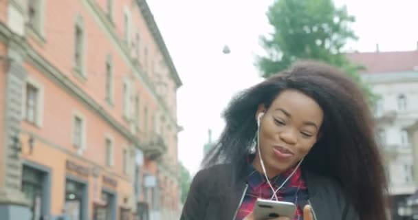 Duygusal mutlu Afrikalı kız sohbet ediyor, cep telefonuyla müzik dinlerken sokakta yürürken mutlu bir şekilde zıplıyor ve saçlarını sallıyor. 4k görüntü. — Stok video
