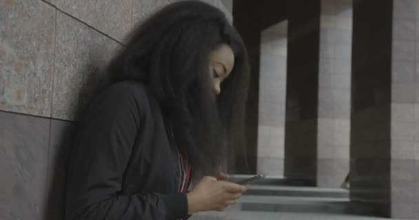 Portrait latéral de la jeune fille africaine magnifique profitant de la musique dans les écouteurs, la danse et le texte via les téléphones mobiles tout en s'appuyant sur le mur en plein air. — Video