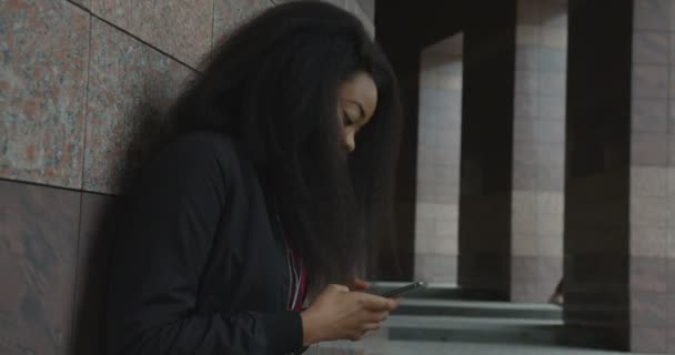 Funny fille africaine énergique avec de beaux cheveux bouclés bavarder et danser tout en écoutant de la musique via les écouteurs près du bâtiment. Images 4k. — Video