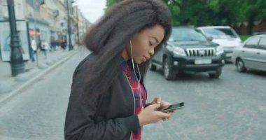 Teknoloji ve İnsanlar. Taşınabilir bağımlılık. Çekici Afrikalı bir kız karşıdan karşıya geçiyor ve cep telefonuyla mesaj atıyor..