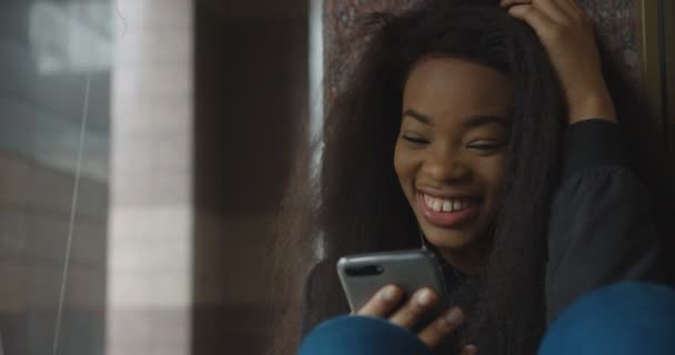 Sourire fille africaine est bavarder et naviguer via le téléphone mobile tout en étant assis sur les escaliers. Portrait de belle femme. Images 4k. — Video