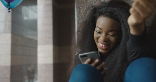 Porträt des hinreißenden afroamerikanischen Mädchens, das lacht, während es SMS schreibt und auf der Treppe mit dem Handy surft. 4k Filmmaterial. Draußen. — Stockvideo