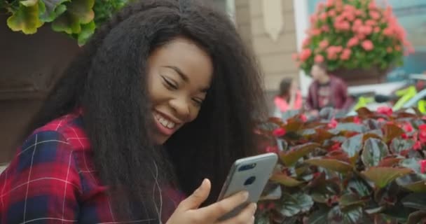 Seite an Seite ein Porträt. Stilvolle glückliche junge afrikanische Mädchen ist SMS über das Handy, während die Musik im Kopfhörer genießen im Freien in Café. — Stockvideo