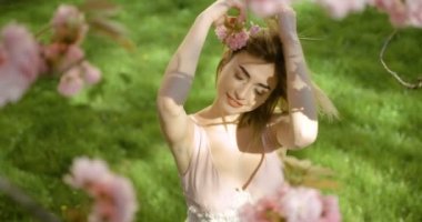 Çayırdaki sakura çiçekleriyle güzel gülüşlü, poz veren ve yüzüne dokunan çekici sarışın kıza tepeden bak. 4k görüntü.
