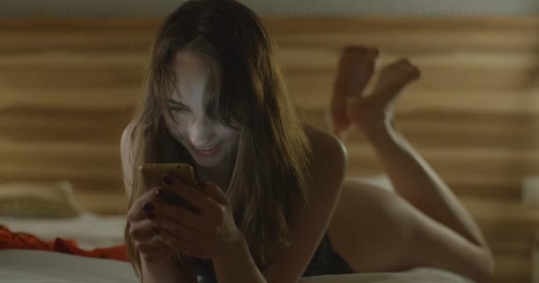 Ritratto della bella bruna in lingerie nera di pizzo sorridente e sms, chatta e naviga tramite il telefono cellulare mentre è sdraiato sul letto. 4k filmati. — Video Stock