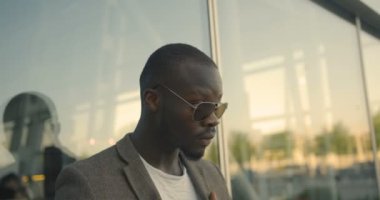 Cam binanın yanındaki şık ceketini düzelten güneş gözlüklü yakışıklı Afro-Amerikalı adamın portresi. 4k görüntü.
