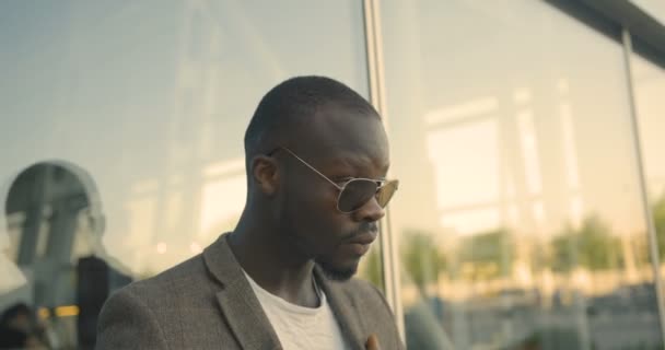 モダンなサングラスのスタイリッシュな自信に満ちたアフリカ人男性は、ガラスの建物の近くで彼のジャケットを修正しています。4k映像. — ストック動画