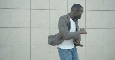 Beyaz duvarın arkasında yürürken şık giyinmiş ve güneş gözlüğü takmış komik, gülümseyen Afrikalı adam dans ediyor. 4k görüntü.