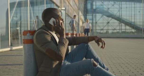 Side portret van de zelfverzekerde stijlvolle Afrikaanse man emotioneel praten en het maken van gebaren tijdens het praten via de mobiele telefoon, zittend op het asfalt en leunend op de wegpaal. — Stockvideo