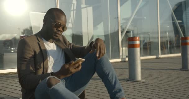 Attraente uomo africano moderno con occhiali da sole sta parlando tramite il telefono cellulare mentre seduto sulla strada e appoggiato sul palo. 4k filmati. — Video Stock