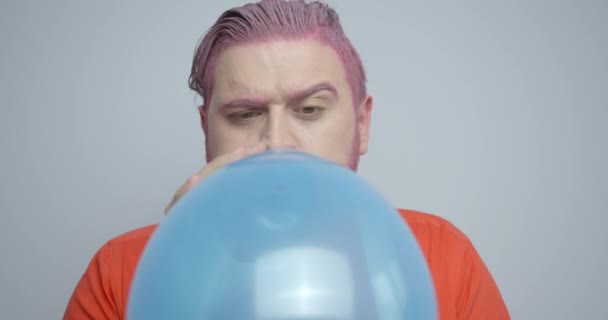 Retrato emocional do homem com cabelo rosa e barba soprando o balão azul e está aparecendo no fundo branco. Filmagem 4k. — Vídeo de Stock