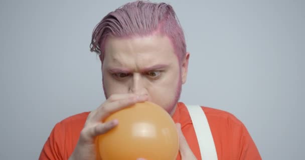 Porträt des rosafarbenen Kopfmenschen, der den orangefarbenen Ballon in die Luft sprengt. Seine Reaktion auf den Knall. Er reinigt das Ohr mit dem Finger. 4k Filmmaterial. — Stockvideo