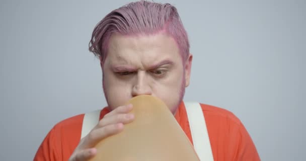 Růžová hlava mladý hipster muž fouká do oranžového balónu, to praskne a tahá uši. Umyje to ucho prstem. Záběry 4k. — Stock video