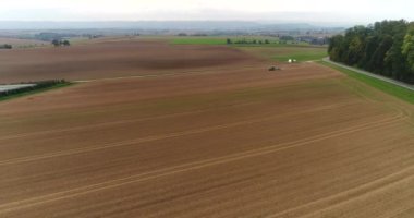 Almanya 'da yoğun yeşil ormanlarla çevrili güzel hasat tarlaları üzerinde hava uçuşları. Çalışan tarım makinesinin üzerinde uçan helikopter..