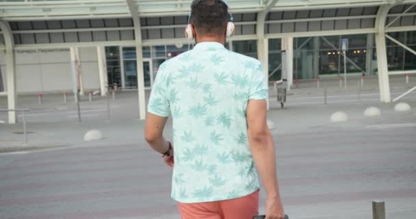 Vista trasera del turista africano en auriculares tirando de su equipaje mientras camina hasta el aeropuerto moderno. Imágenes de 4k. — Vídeos de Stock