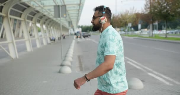 Jovem atraente turista africano em heaphones está caminhando para o aeroporto para um voo. Ele está a puxar a bagagem. 4k. — Vídeo de Stock