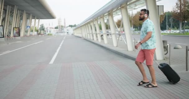 Jovem turista africano bonito em t-shirt, shorts e fones de ouvido está puxando sua bagagem enquanto caminha para o terminal do aeroporto. Vista lateral de comprimento total em 4k. — Vídeo de Stock