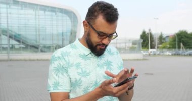 Gözlüklü çekici Afrikalı adam cam binanın yanındaki cep telefonunda geziniyor ve mesajlaşıyor. Döner görüş. 4k görüntü.