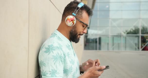 Güneş gözlüklü genç Afrikalı adamın yan portresi kulaklıkla müzik dinliyor ve havaalanı terminali yakınında cep telefonuyla sohbet ediyor. 4k görüntü. — Stok video