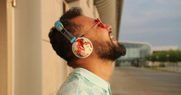 Розслаблений афроамериканський чоловік у стильних рожевих сонцезахисних окулярах та навушниках сміється біля терміналу аеропорту. 4k кадри. Бічний портрет . — стокове відео