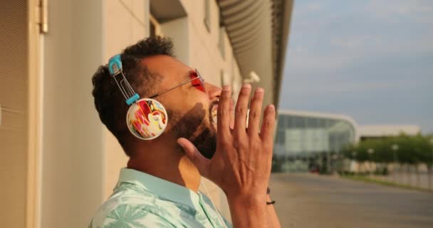 Συναισθηματικό πορτραίτο του νεαρού χαρούμενου Αφρικανού με ροζ γυαλιά ηλίου και ακουστικά που γελάει κοντά στο αεροδρόμιο. 4k πλάνα. — Αρχείο Βίντεο