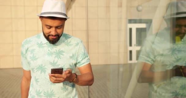 《人物与科技》。一个穿着T恤衫，头戴帽子的快乐的非洲男人的画像，一边聊天，一边在手机上浏览，然后把他的手提箱拖到机场。4k. — 图库视频影像