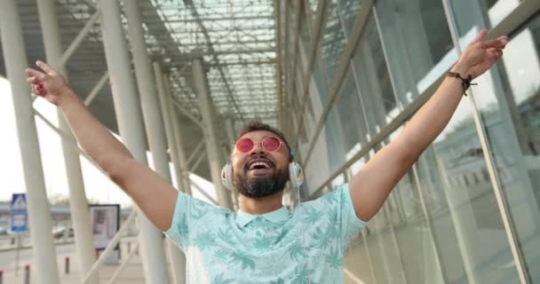 Πορτρέτο του χαρούμενου συναισθηματικού Αφρικανού με τα πολυτελή ροζ γυαλιά ηλίου που ακούει μουσική στα ακουστικά και σηκώνει τα χέρια ψηλά. 4 χιλιάδες πουλάρια. Τοποθεσία αεροδρομίου. — Αρχείο Βίντεο