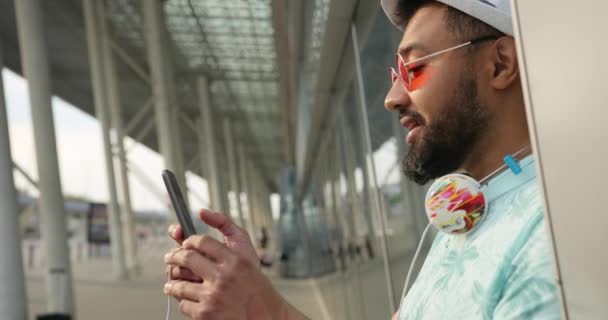 Okouzlující mladý Afričan v klobouku, tričku a slunečních brýlích si povídá a prohlíží si mobilní telefon, zatímco čeká na svůj let poblíž letiště. Záběry 4k. — Stock video