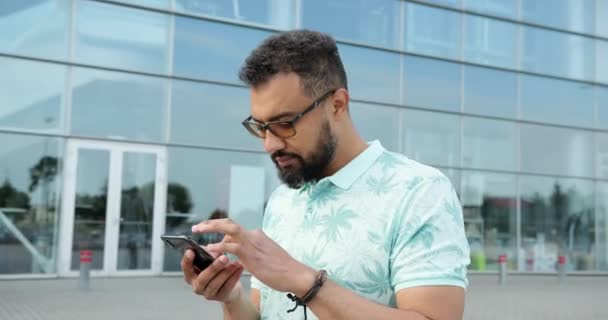 在现代玻璃建筑附近，一个戴着眼镜的英俊笑脸的非洲男人一边聊天一边在手机上浏览，这让人眼花缭乱。4k. — 图库视频影像