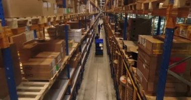 Araba fabrikası deposunun içinde. Bir grup işçi, araç gereçleri, elektrik kabloları ve kablolarla raflarda yürüyorlar. İçlerinden biri palet kamyonunu kullanıyor..