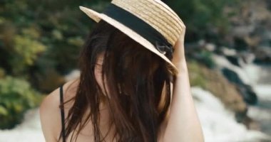 Saman şapkalı, şelalenin önünde duran ve kameraya gülümseyen sevimli genç kadına tepeden bak. Yaz mesleği