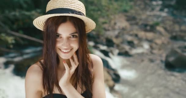 Guarda dall'alto l'adorabile giovane donna con un cappello di fieno in piedi davanti a una cascata e sorridente direttamente alla fotocamera — Video Stock