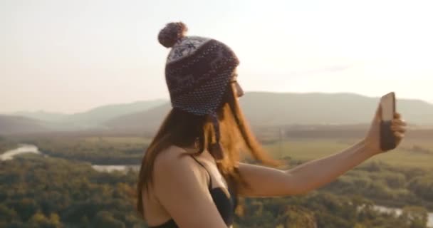 Γοητευτική νεαρή hipster κορίτσι πηδά με ένα smartphone και χαμογελά μιλώντας σε μια βιντεοκλήση. Ορεινή καλοκαιρινή κλίση — Αρχείο Βίντεο