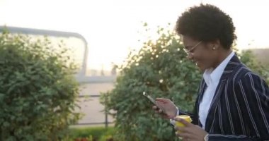 4 bin. Ağır çekim. Olumlu duygular. Şık giyinmiş Afro-Amerikalı iş kadını elinde bir fincan kahveyle yürüyüp akıllı telefonundan bir şeyler okuyor.