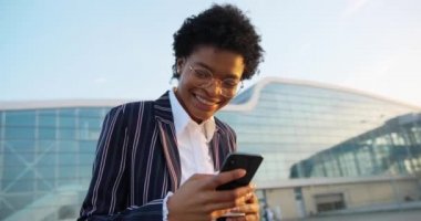 4 bin. Ağır çekim. Çekici Afrikalı Amerikalı kadın telefonunda bir şey okurken gülümsüyor ve akşam güneşinin altında modern binanın önünde dikilip kahve içiyor.